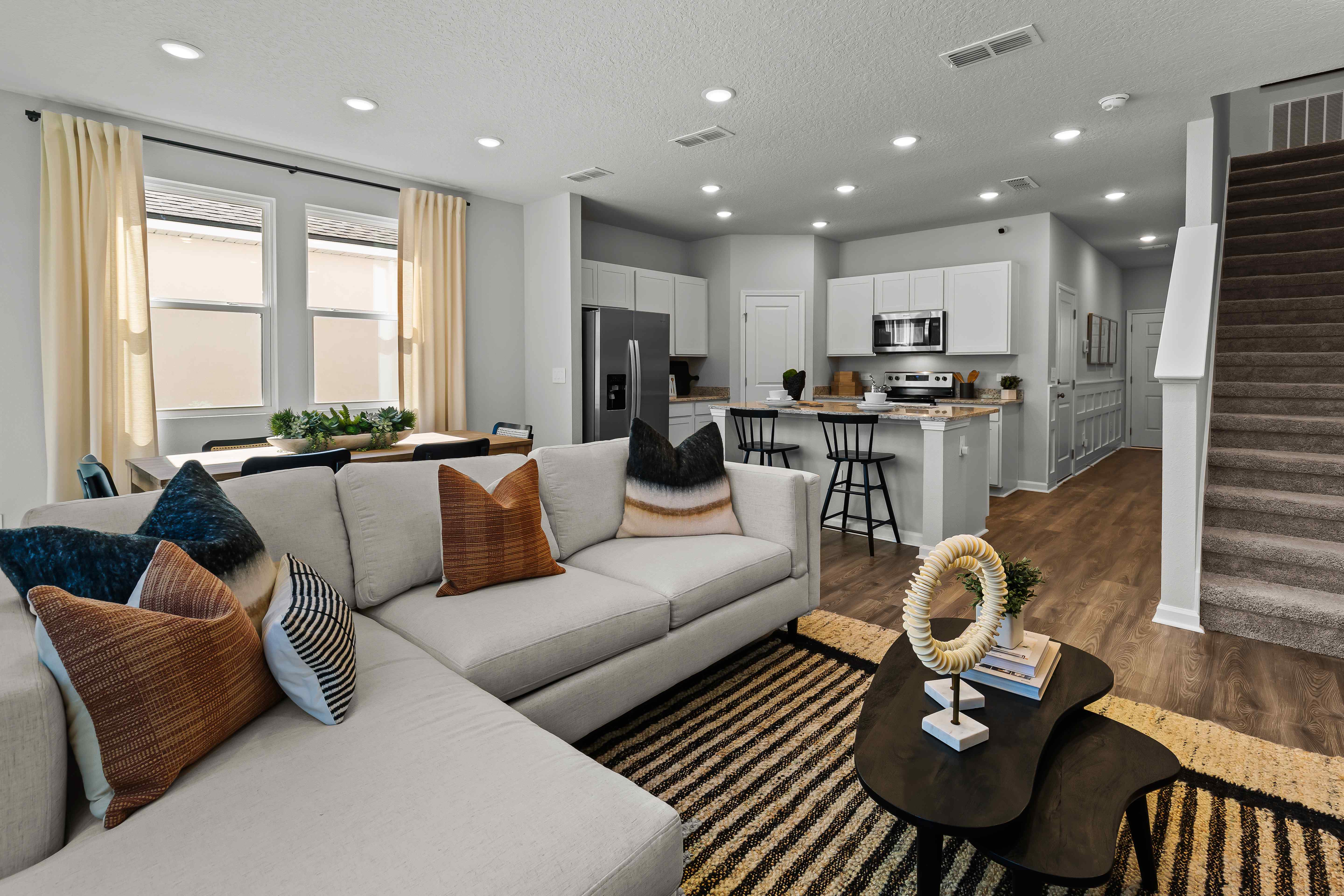 Davor Model Paired Home Living with Cream Couch Looking over Kitchen with White Cabinets and White Staircase at The Landings at Pecan Park